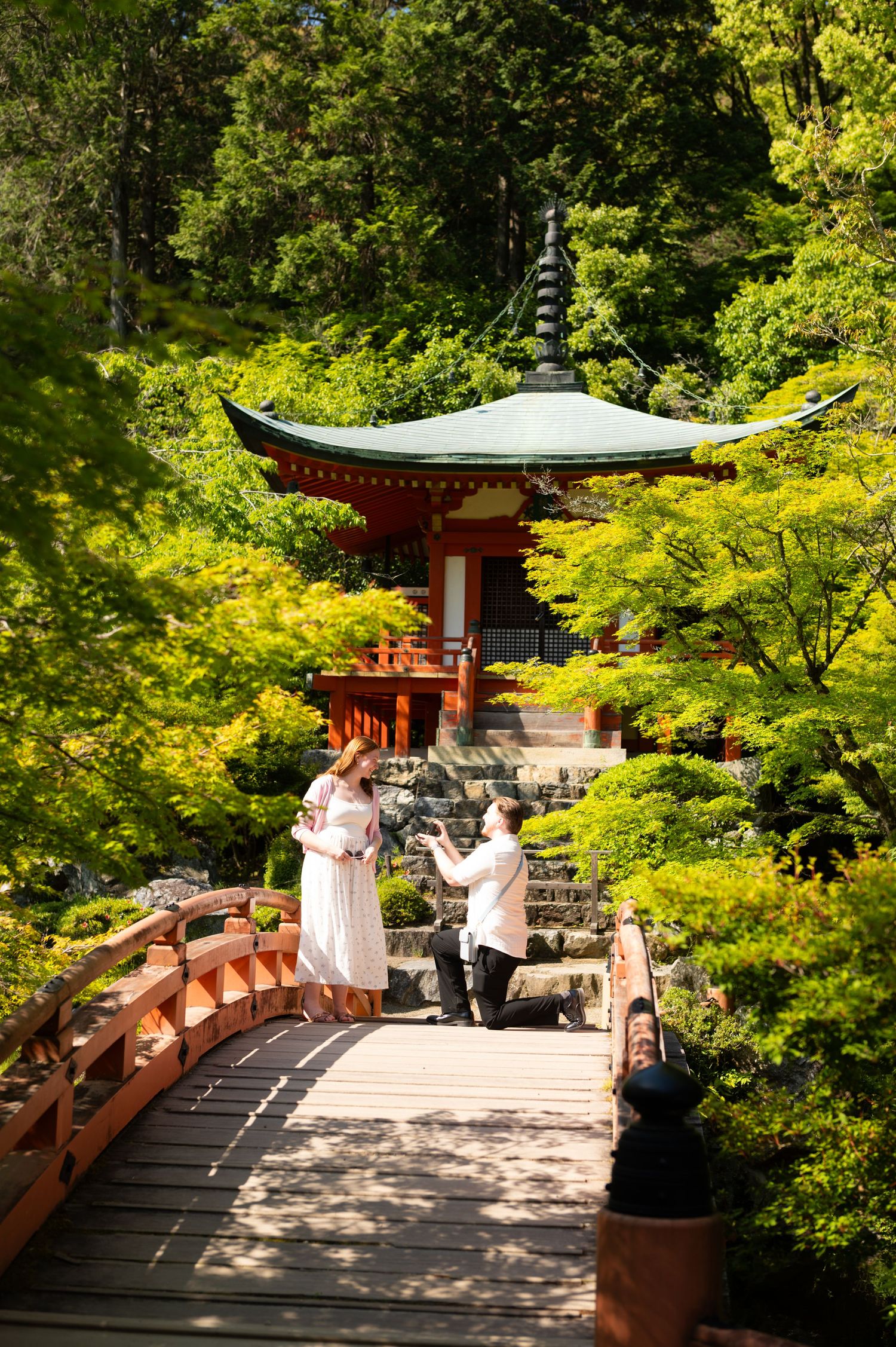 Man kneeling on one knee proposing to a woman in a white dress near a red wooden bridge and pagoda surrounded by lush green trees.