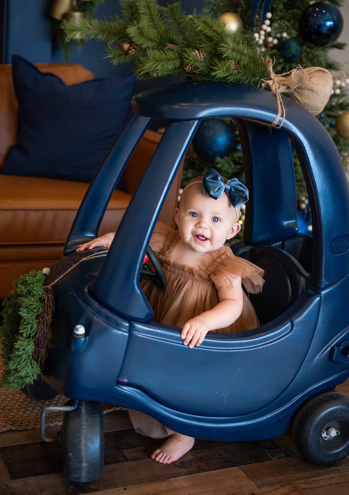 Smiling baby girl in a tan tulle dress and blue velvet bow sits in a navy toy car with Christmas tree and evergreen garland decor.