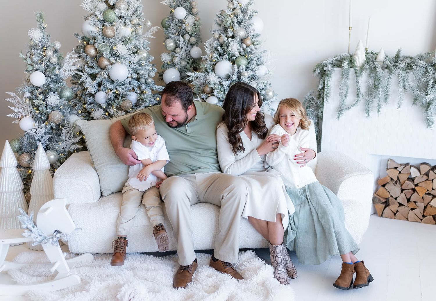 Family of four sitting on a white sofa in a bright Christmas photoshoot with frosted trees, ornaments, and a fur rug.