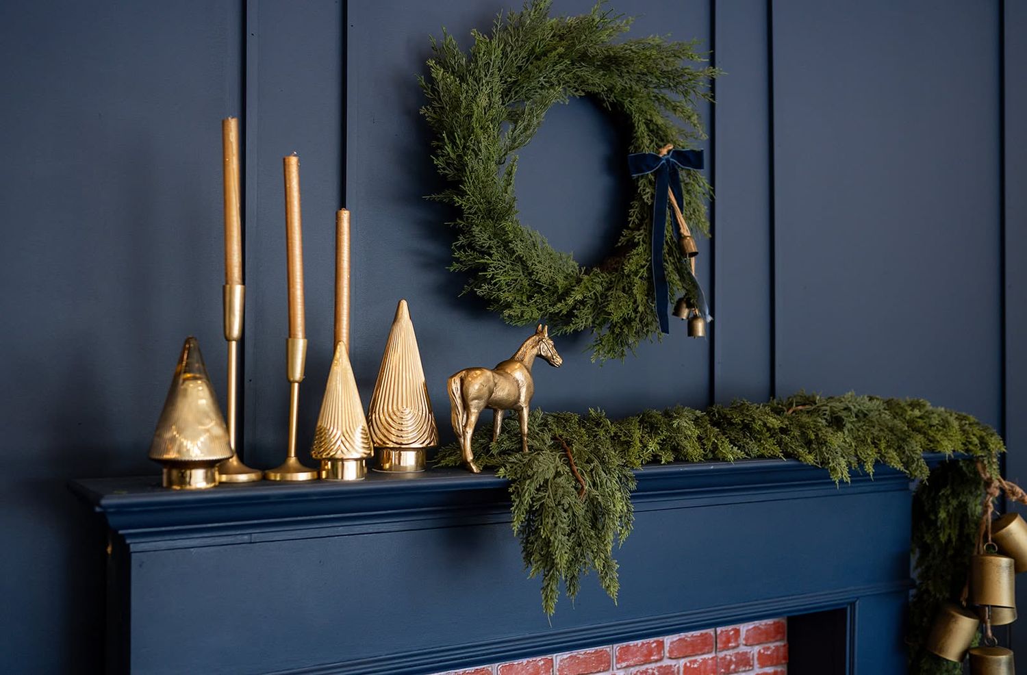 Navy blue fireplace mantel decorated with a evergreen wreath, garland, gold candlesticks, brass trees and a golden horse figurine.