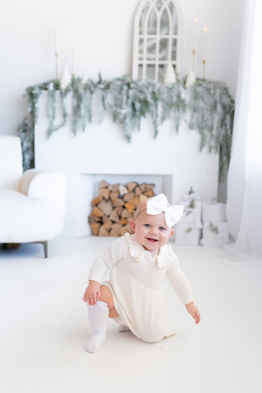 Smiling baby in a cream outfit and white bow crouches on a white floor in front of a decorated fireplace with stacked birch logs.