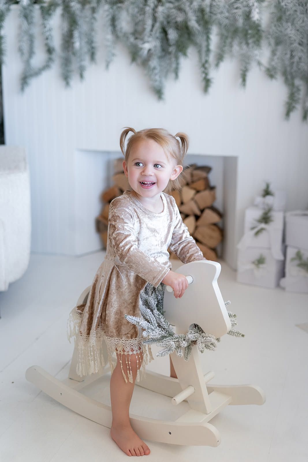 Smiling toddler girl in a sparkly gold dress riding a white wooden rocking horse beside a fireplace with stacked logs and winter greenery.