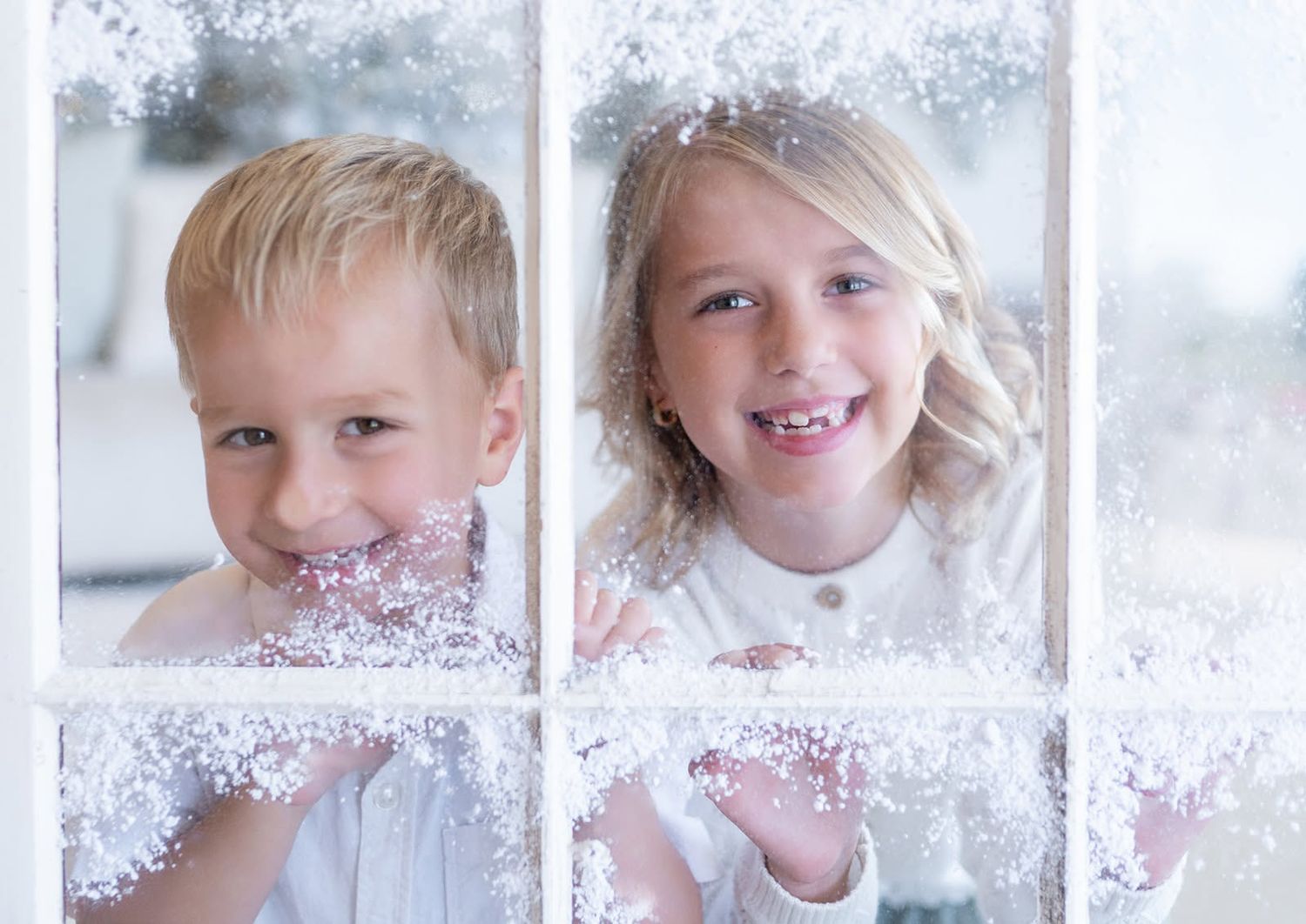 Two smiling children, a boy and a girl, peek through a frosted window pane decorated with snowflakes and winter frost.