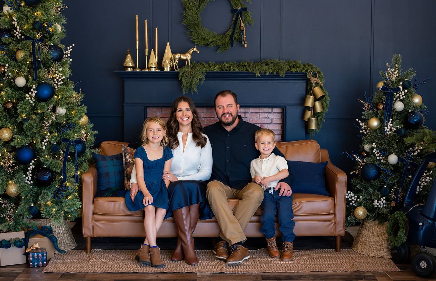 Family of four in coordinating outfits sits on a tan leather sofa in front of a navy fireplace with decorated Christmas trees.