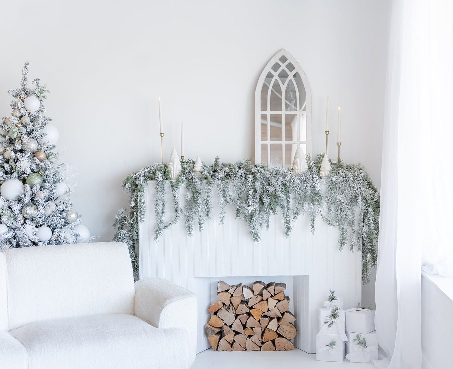 White Christmas living room with a frosted garland mantel, stacked firewood, arched mirror, flocked tree and a cozy white armchair.