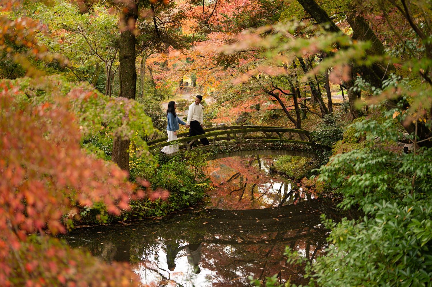 Two people walk across a wooden bridge over a calm pond in a Japanese garden surrounded by vibrant red and orange autumn foliage.