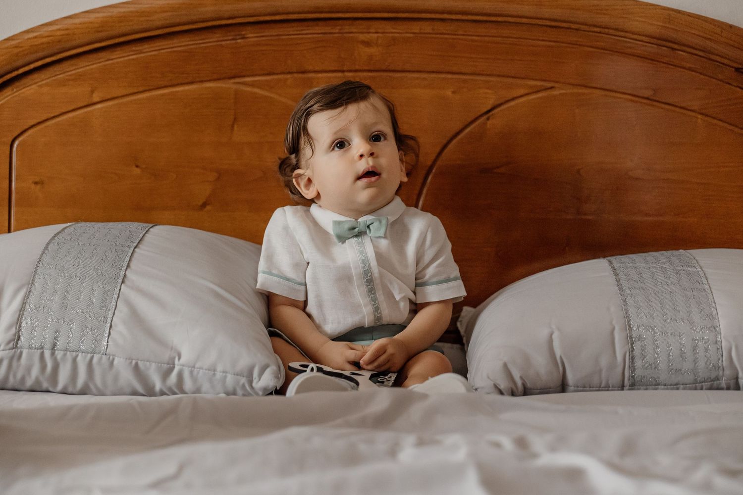 Adorable baby boy in white shirt and mint shorts sitting on a bed, clapping hands and smiling.
