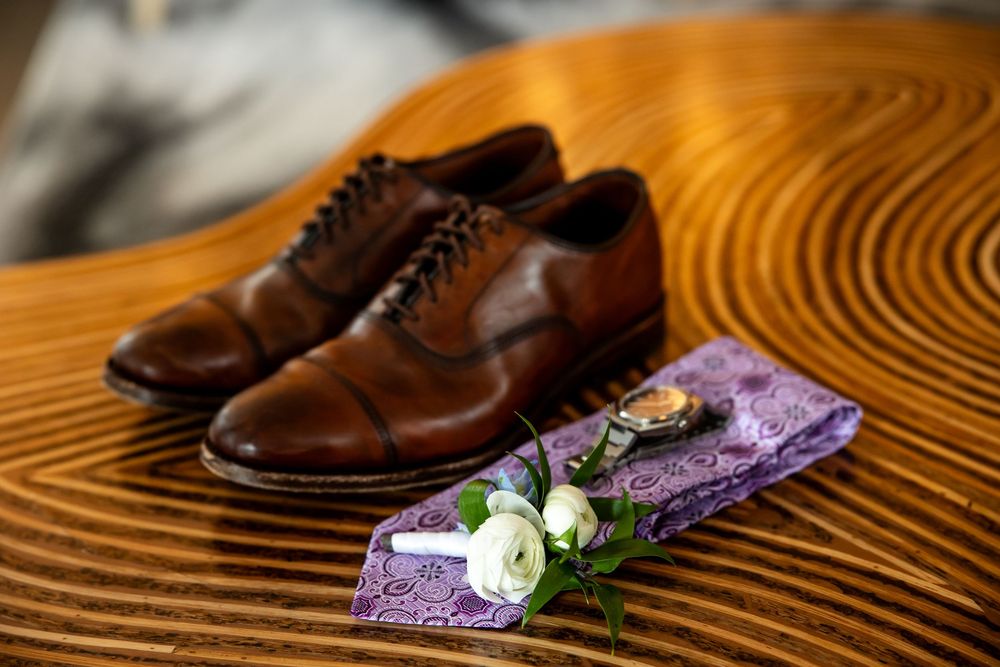 Brown leather oxford shoes with purple paisley tie, boutonniere, and cufflinks on wooden surface for wedding.