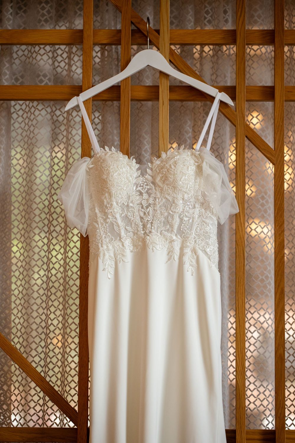 Elegant white lace wedding dress with flutter sleeves hanging on a white hanger against a wooden lattice backdrop.