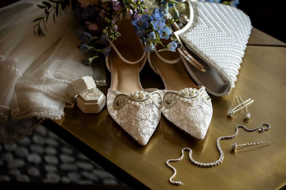 Elegant bridal flat shoes with lace detail displayed alongside pearl clutch, jewelry, and bouquet on gold surface.