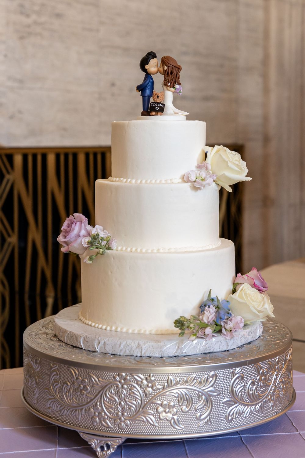 Three-tier white wedding cake with floral decorations and bride and groom cake topper on silver embossed stand.
