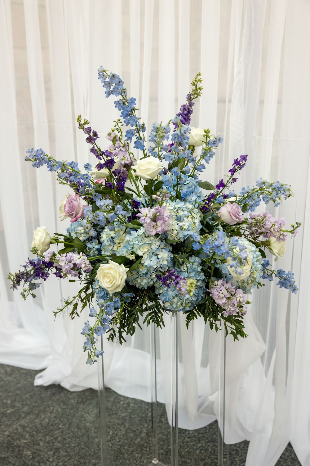 Elegant floral arrangement featuring blue hydrangeas, white roses, purple delphiniums, and pink blooms on a glass stand.
