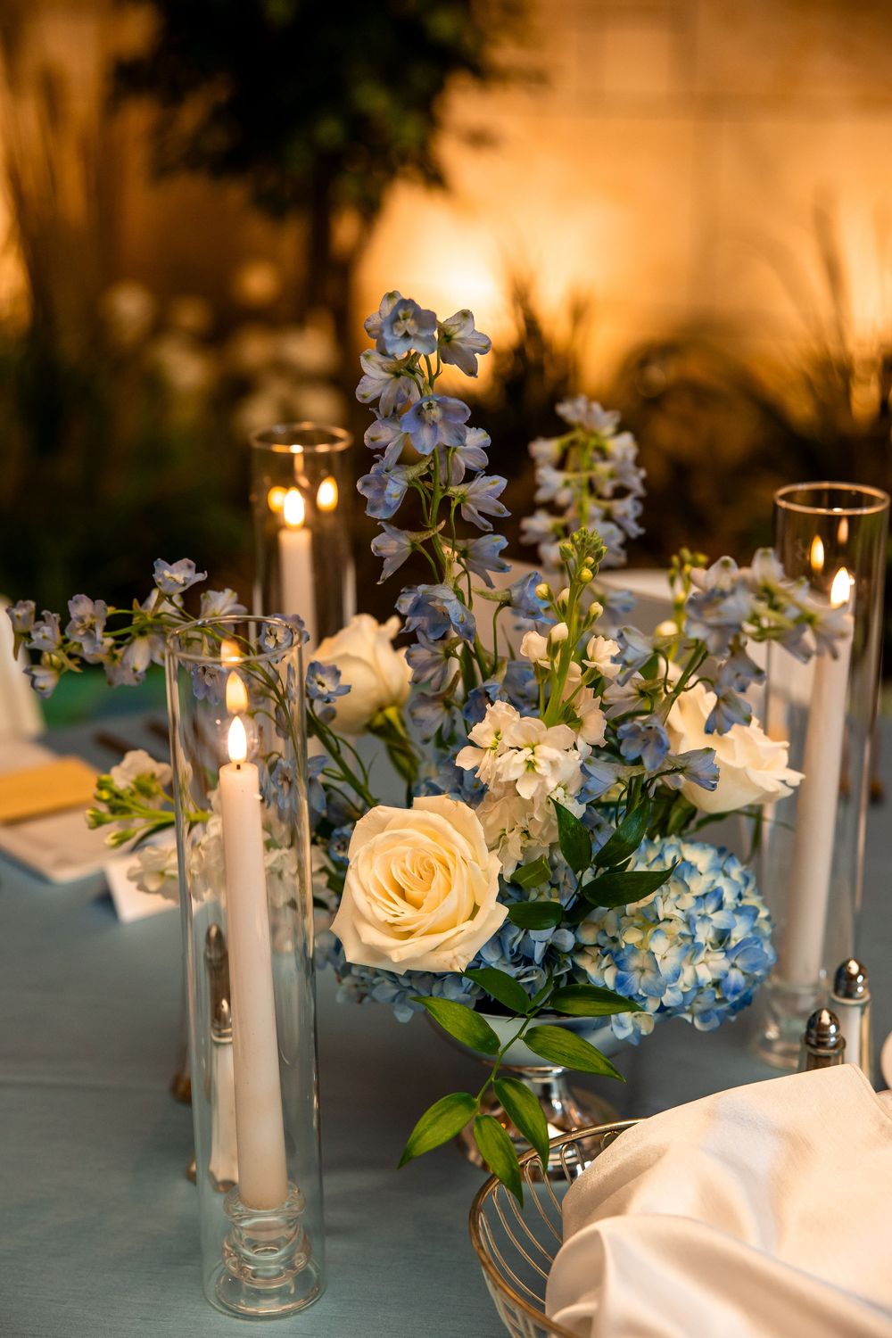 Elegant wedding table centerpiece with blue hydrangeas, yellow roses, delphinium, and tall glass candle holders on a light blue tablecloth.