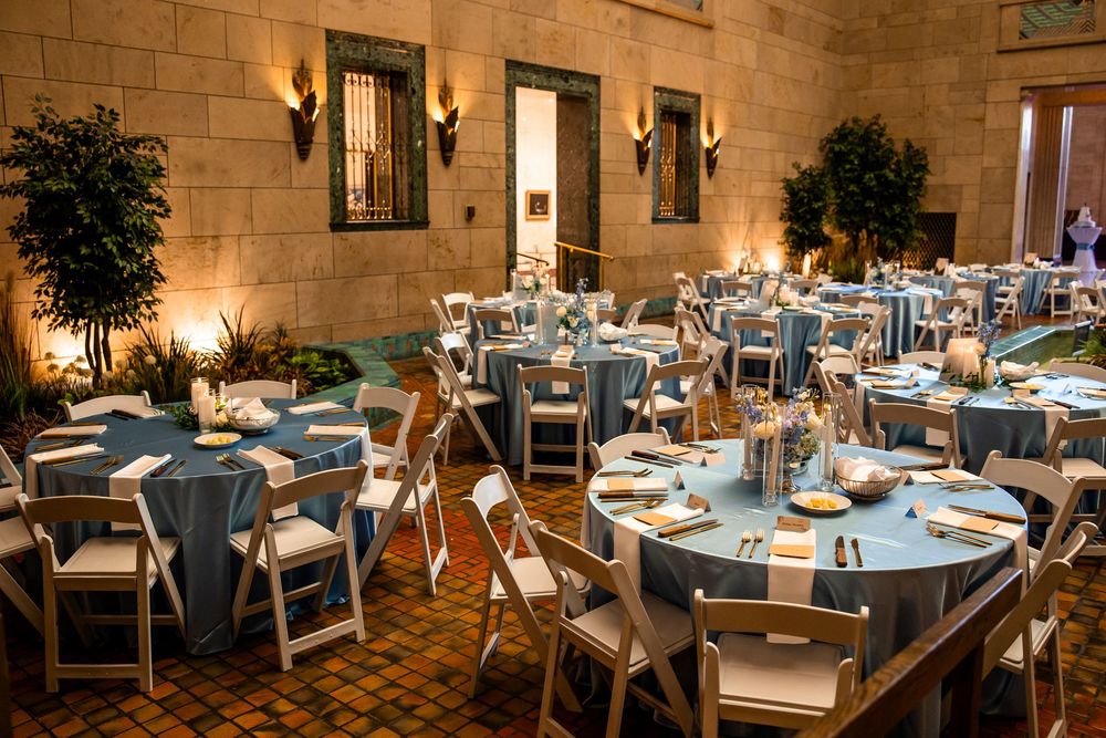Elegant banquet hall with round tables draped in blue linens, white folding chairs, and warm wall sconces in a stone venue.