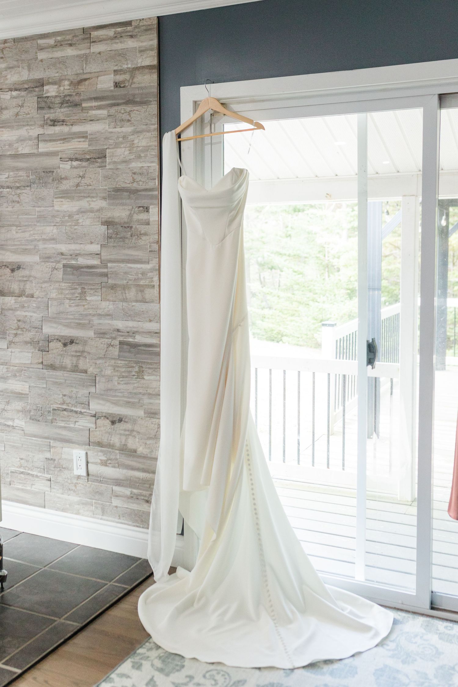 Elegant white wedding dress with a long train hanging on a wooden hanger in front of glass patio doors.