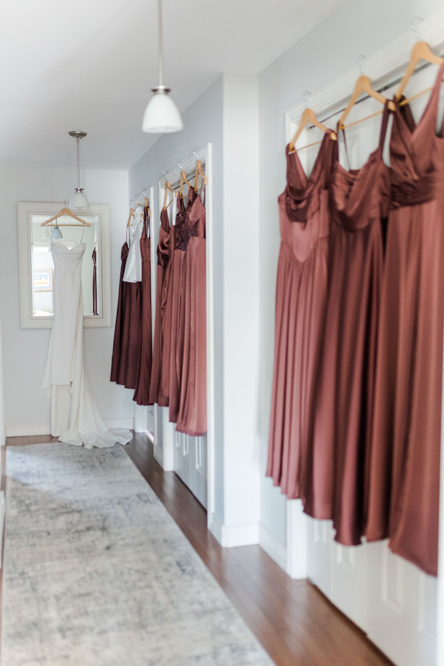Bridesmaids dresses in dusty rose and burgundy hanging on a rack beside a white wedding gown in a bright hallway.