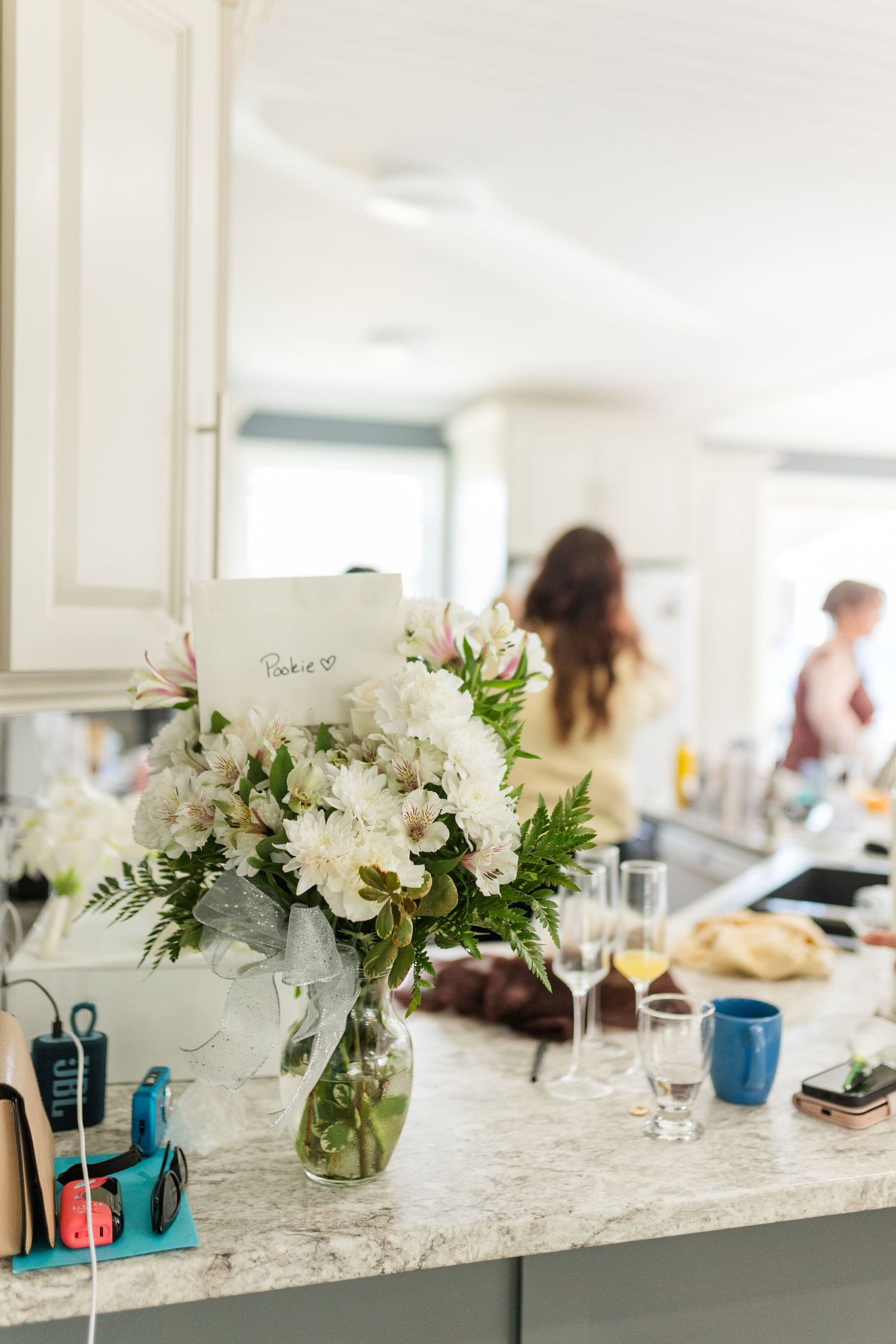 White floral bouquet in glass vase on marble kitchen counter during a casual gathering with guests in background.