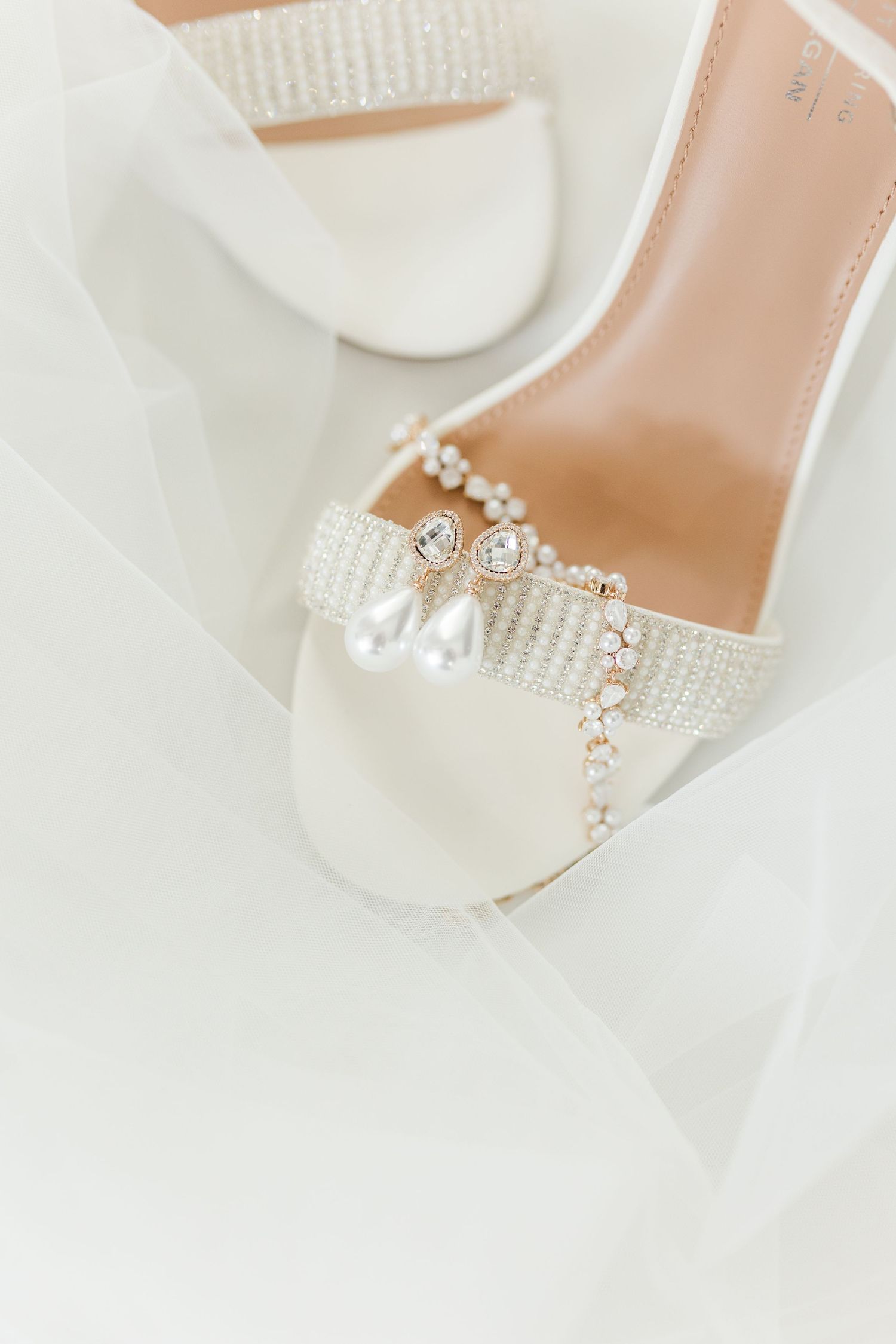 White bridal heels with pearl and crystal embellishments displayed on a wedding veil with matching drop earrings.