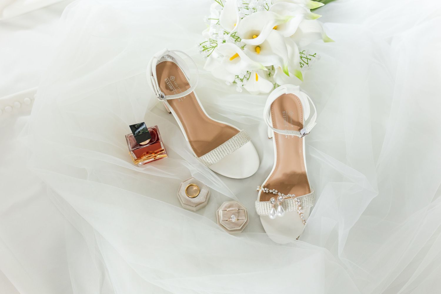 White bridal heeled sandals with ankle strap displayed on tulle with perfume, rings, and white calla lilies.