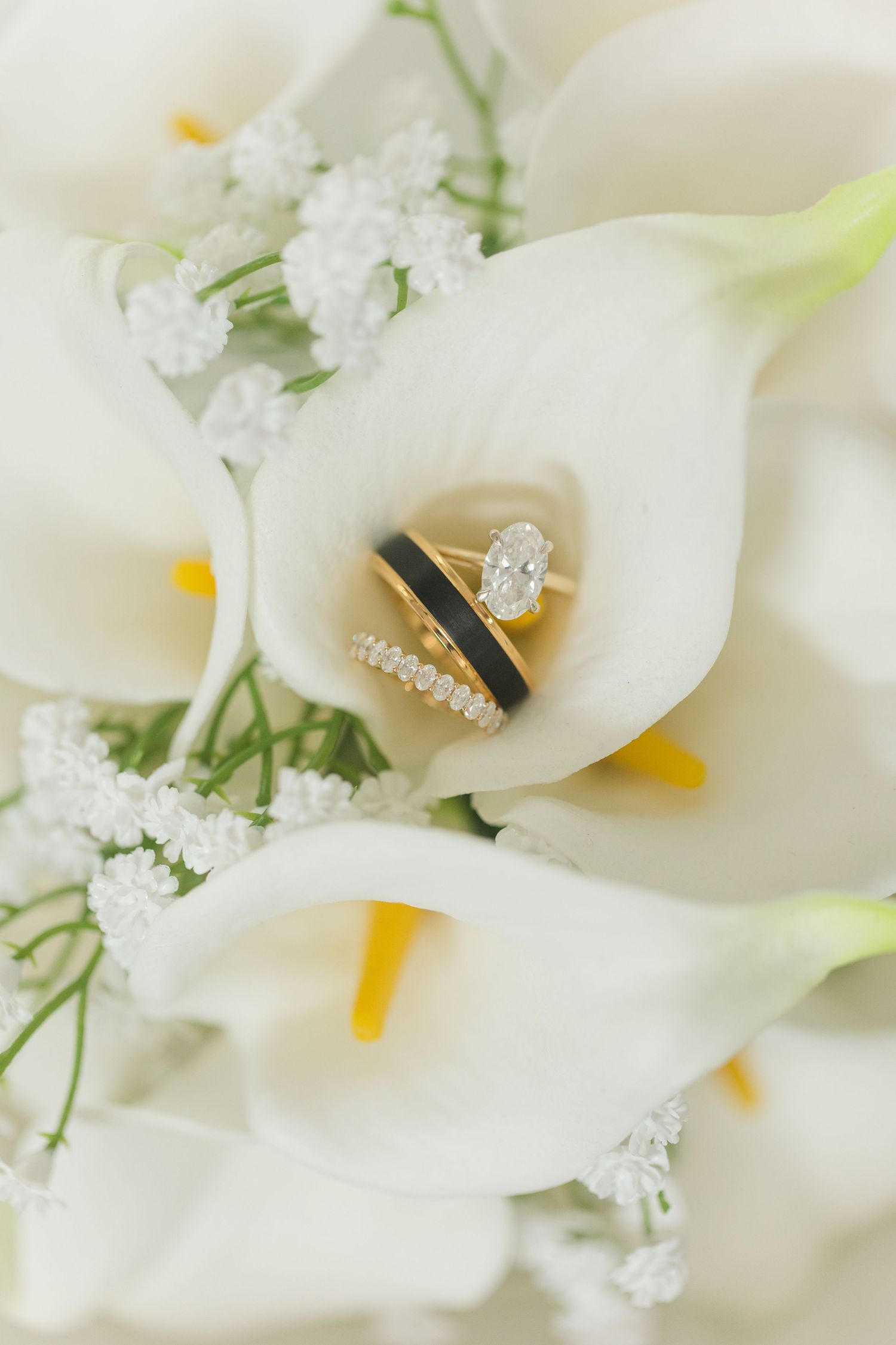 Gold oval diamond engagement ring and black wedding band nestled in white calla lily bouquet with baby's breath.
