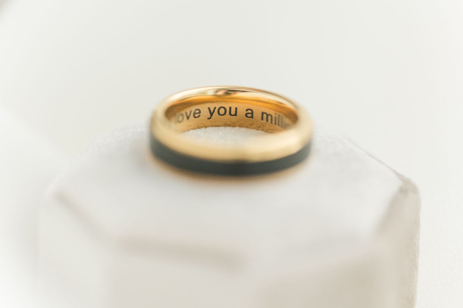 Gold wedding band with engraved message 'love you a mil...' displayed on a white geometric stand.