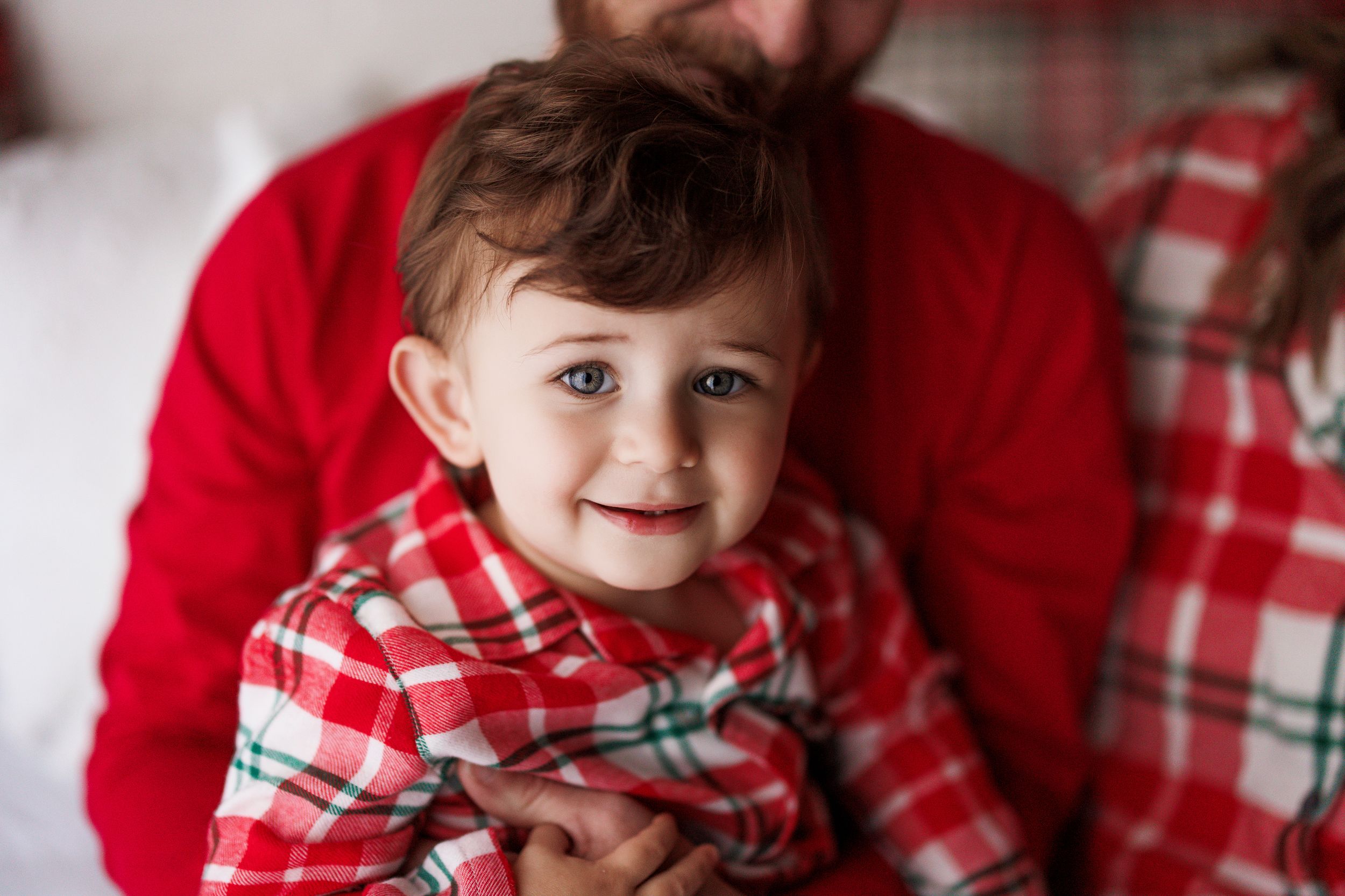 Adorable toddler in red plaid Christmas pajamas smiling while being held by an adult in matching holiday outfit.