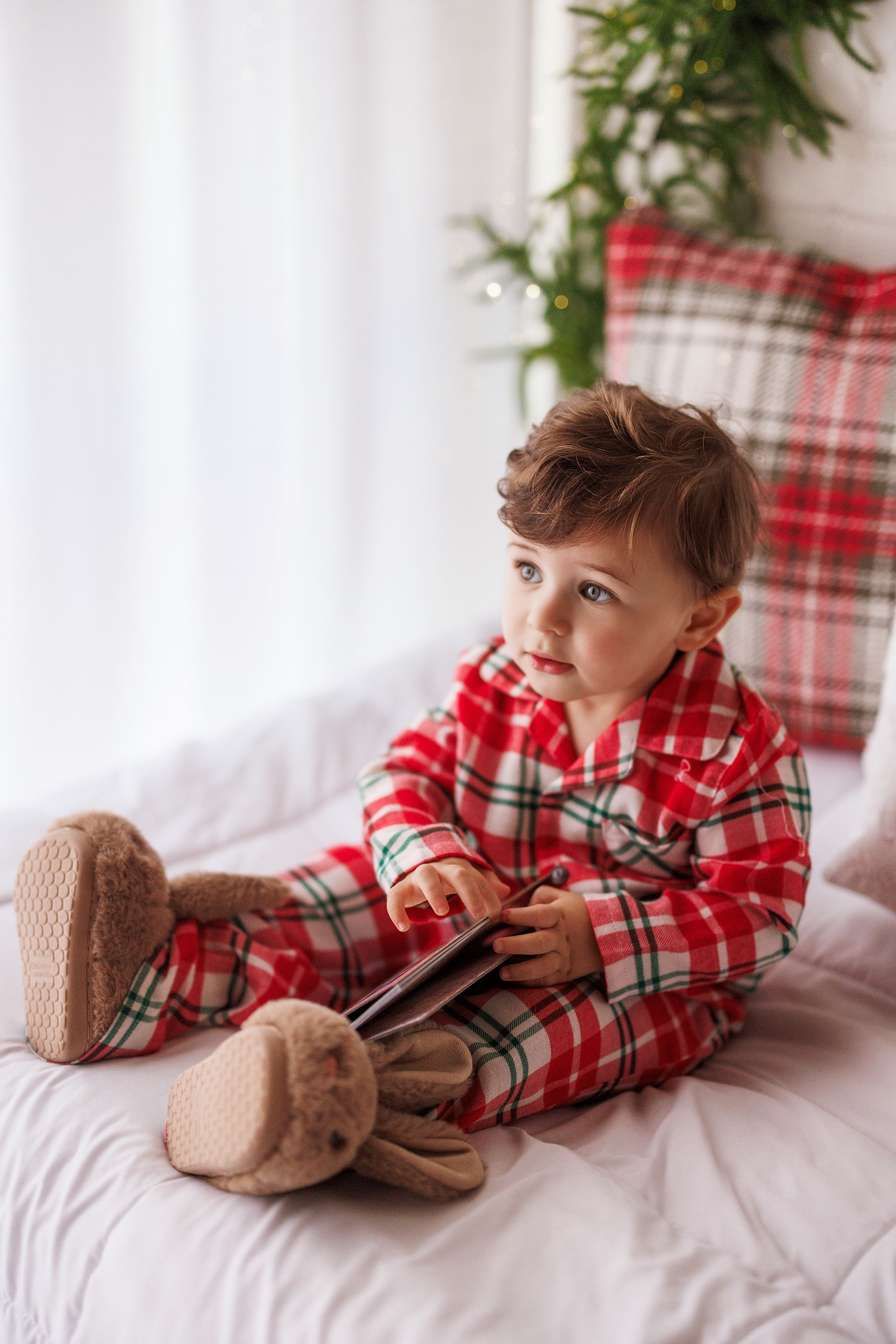 Toddler in red plaid Christmas pajamas and bunny slippers sitting on white bed holding a book.