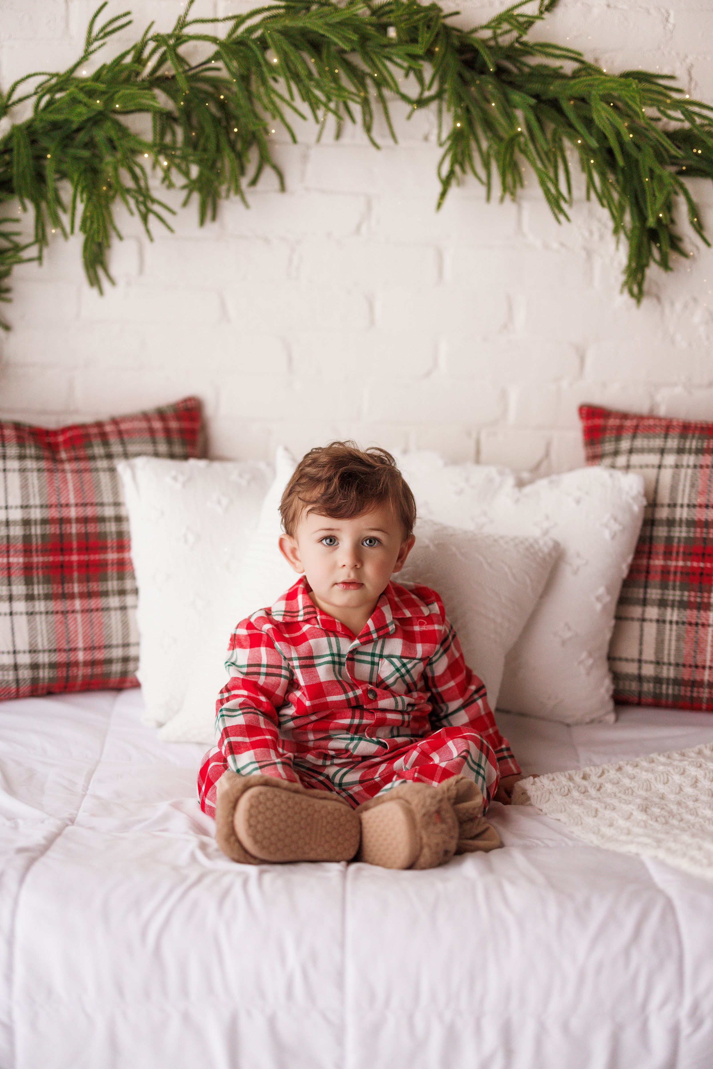 Toddler in red plaid Christmas pajamas sitting on a white bed with plaid pillows and evergreen garland.