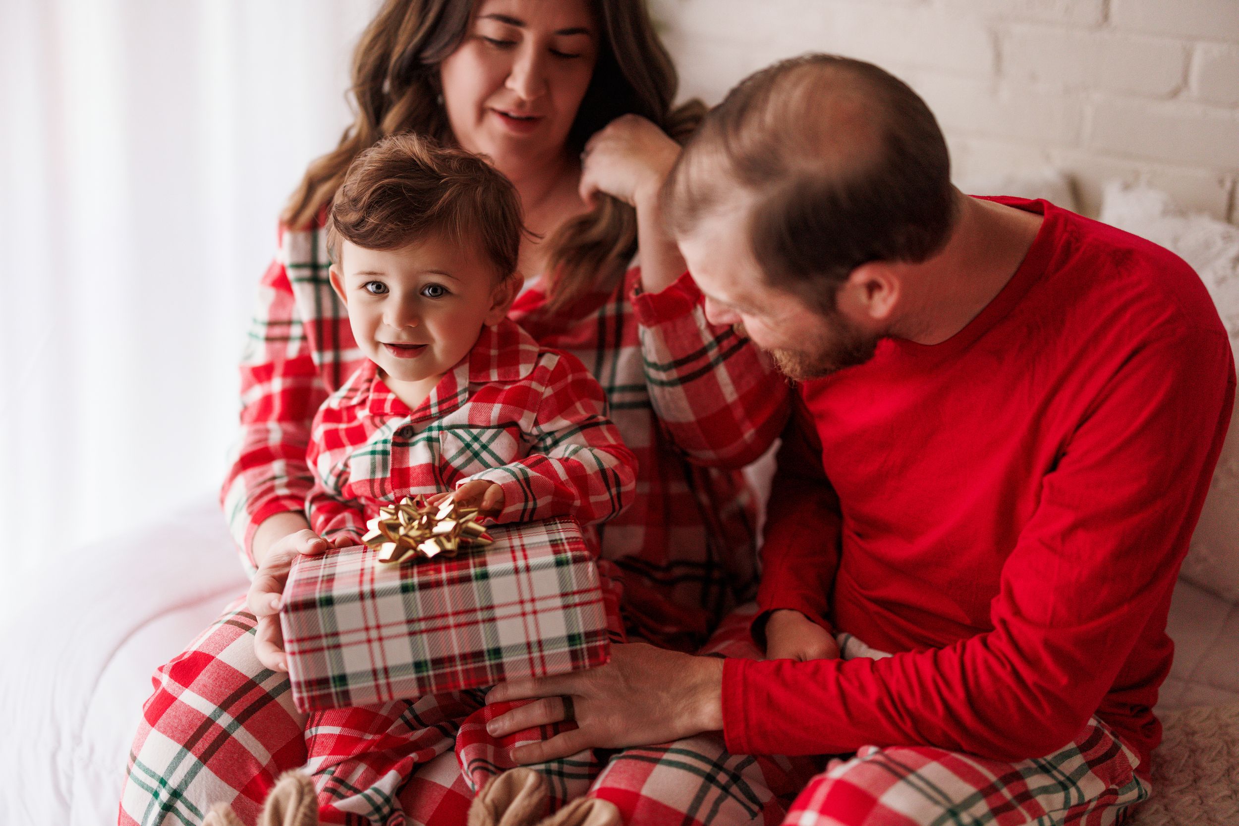 Family in matching Christmas plaid pajamas with toddler holding a wrapped gift with gold bow.