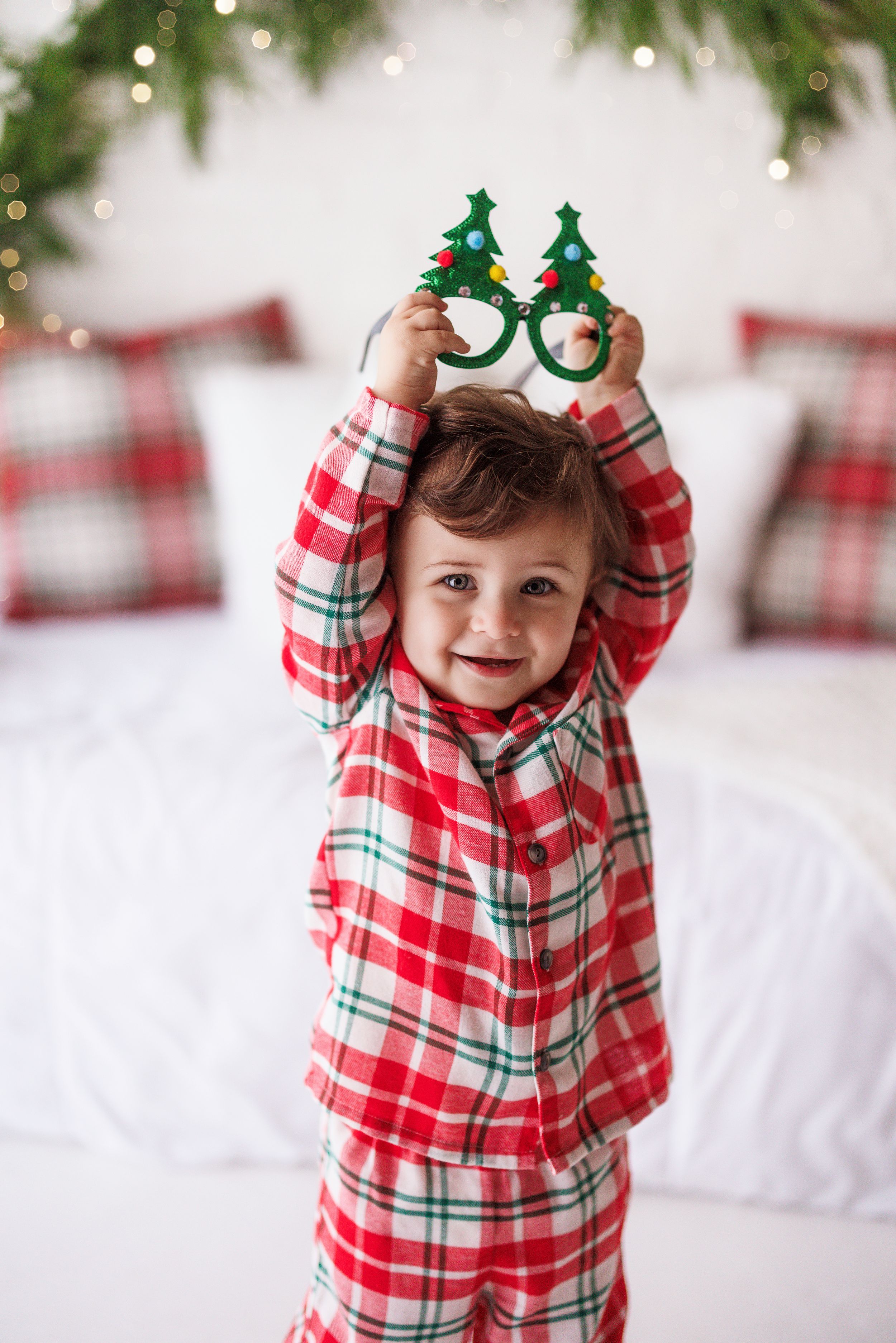 Toddler in red plaid Christmas pajamas holding festive Christmas tree novelty glasses above their head.