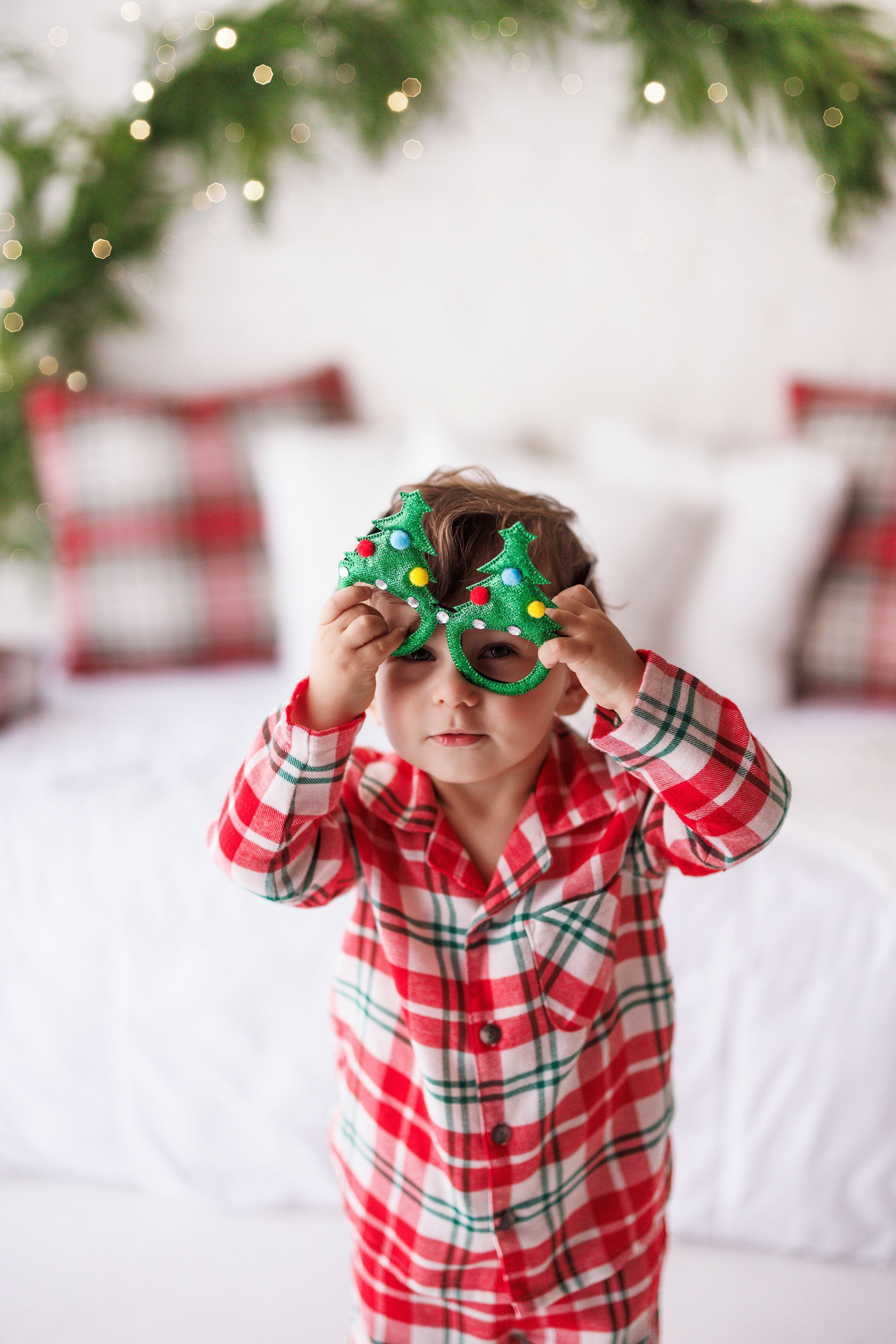 Child in red plaid Christmas pajamas holds festive green tree-shaped cookies over eyes in decorated bedroom.