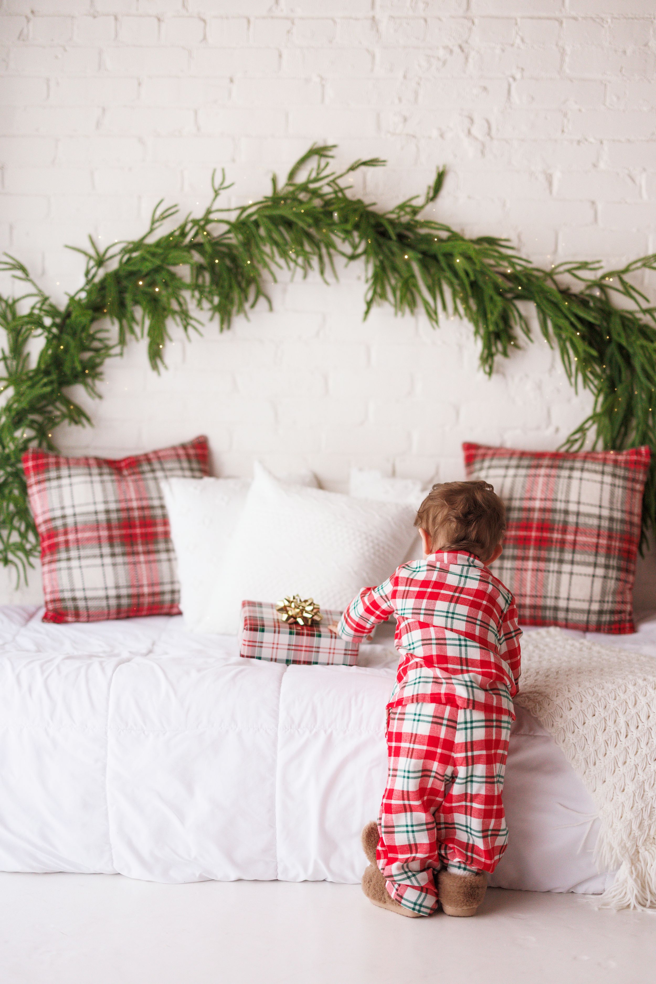 Toddler in plaid Christmas pajamas reaching for a gift on a white bed with holiday garland and tartan pillows.