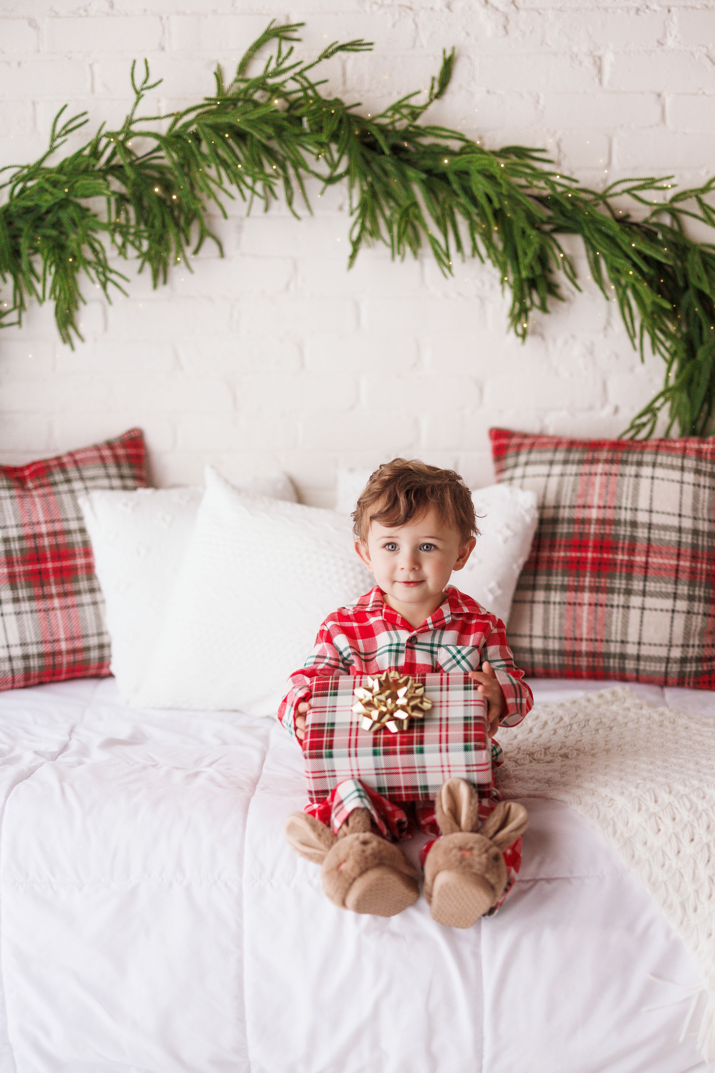 Toddler in plaid Christmas pajamas sitting on bed holding gift with gold bow, surrounded by festive holiday decor.