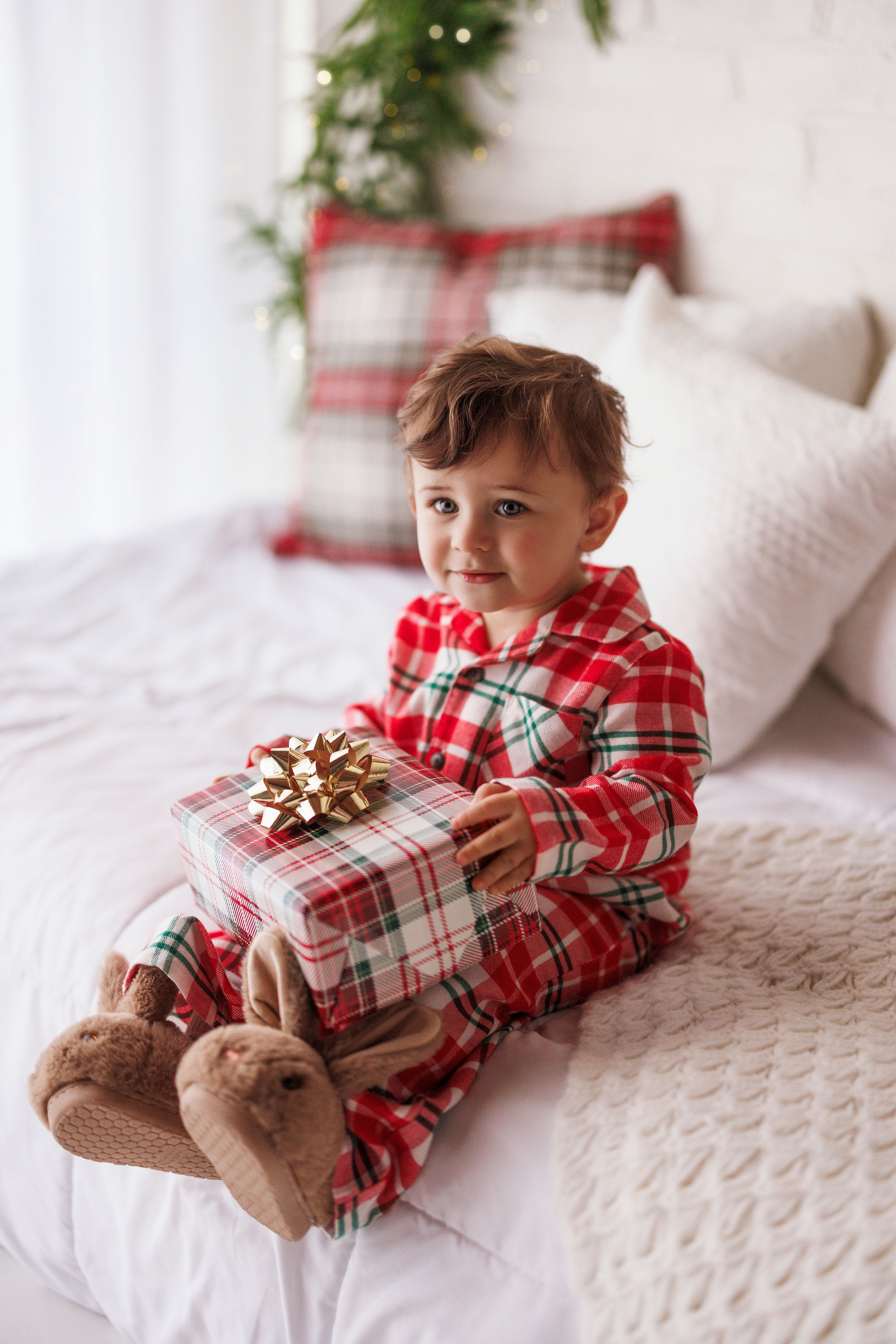 Toddler in red plaid Christmas pajamas sitting on bed holding a gift box with gold bow.