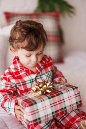 Young child in red plaid pajamas holding a wrapped Christmas gift with a gold bow, sitting on a bed.