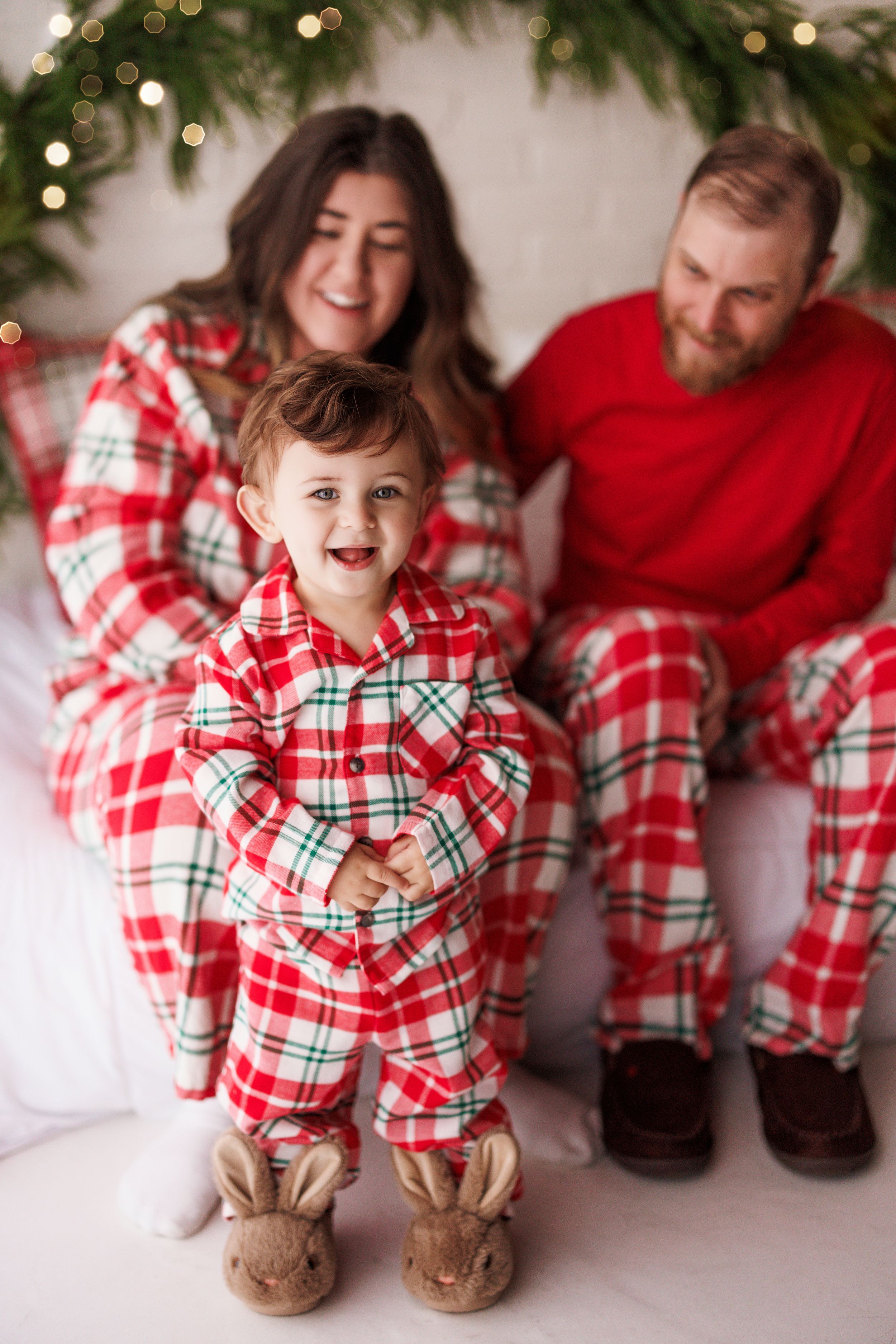Family in matching red plaid Christmas pajamas, toddler standing in bunny slippers smiling, parents seated behind near holiday decorations.