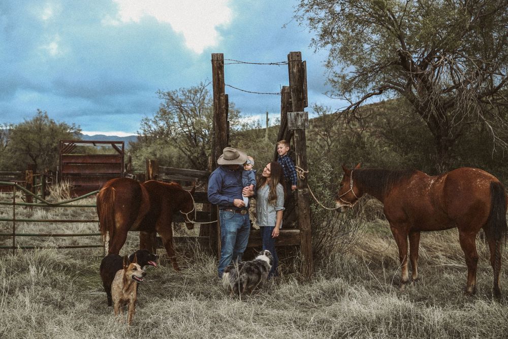 Western family portrait of a man in a cowboy hat, woman and child at a wooden gate with horses and dogs in a rural setting.