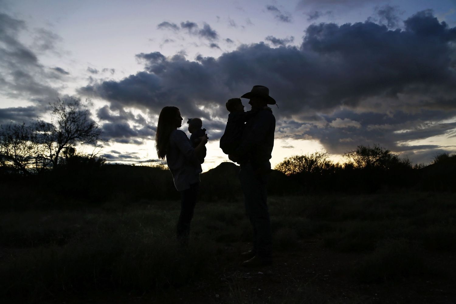Silhouette of a couple holding two small children against a dramatic cloudy sunset sky, the man wearing a cowboy hat.