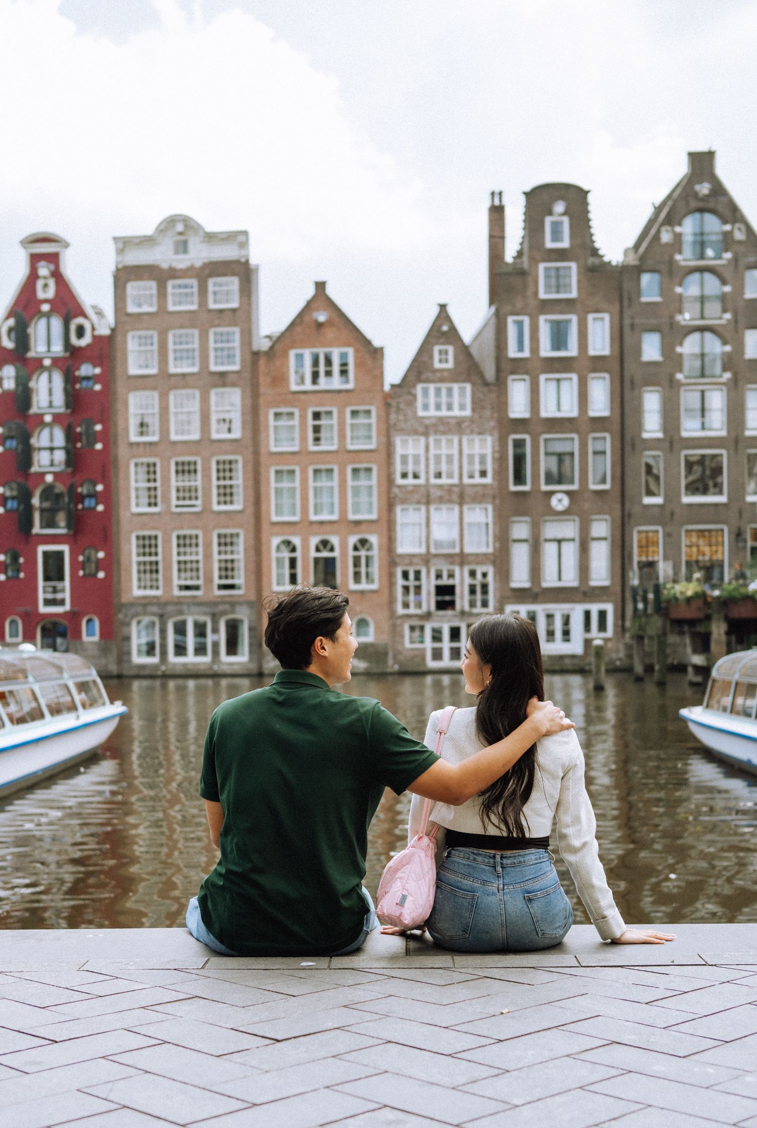 Couple sitting arm in arm on a canal edge in Amsterdam, facing colorful narrow Dutch canal houses near Centraal station.