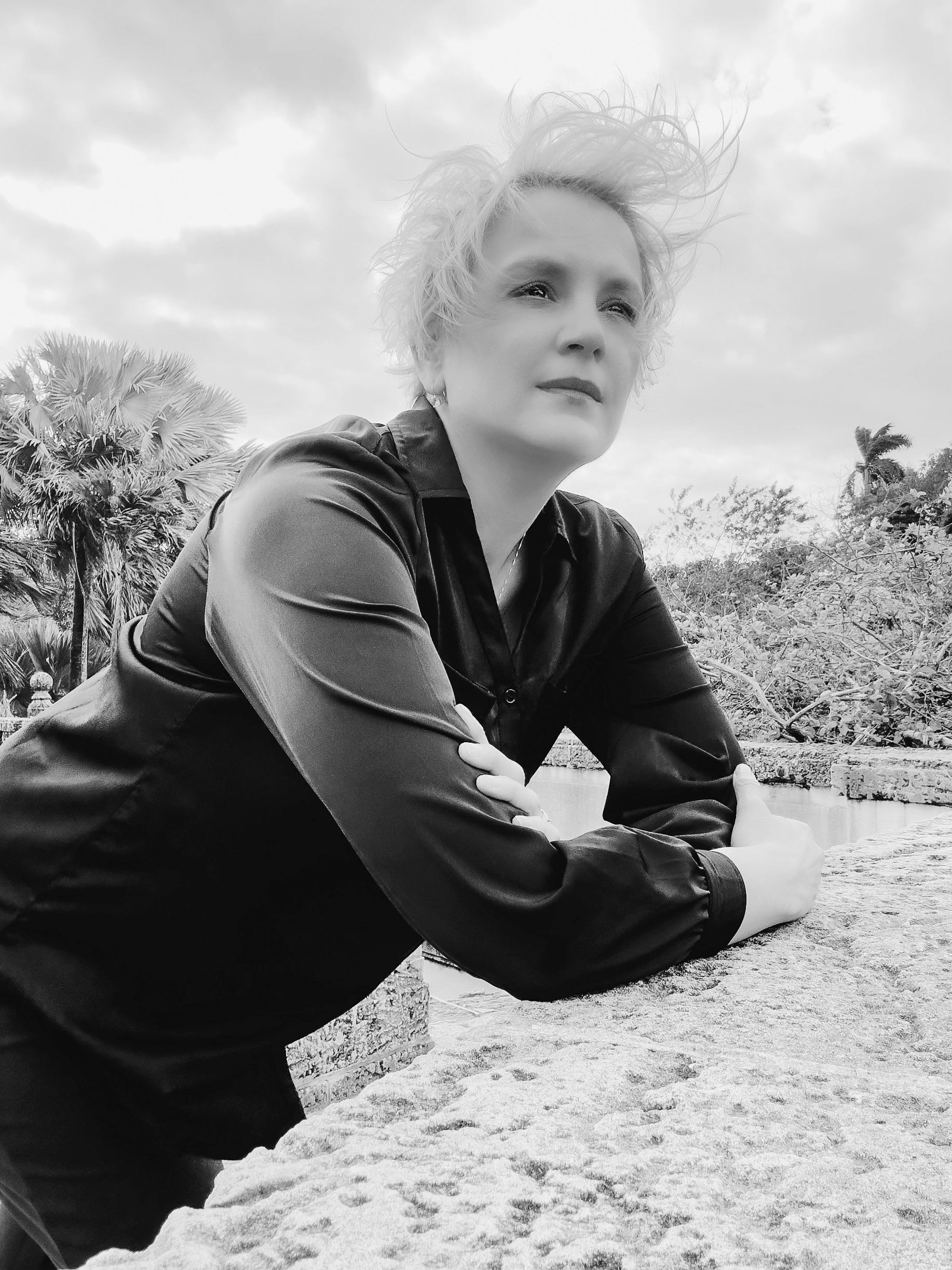 Black and white photo of a blonde person in a black shirt leaning on a stone wall with palm trees behind.