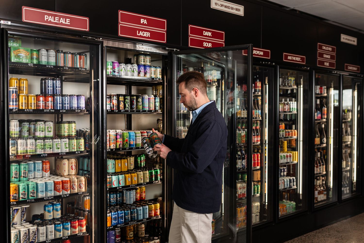 Man in a navy jacket browses a can in front of glass-door coolers stocked with craft beer under Pale Ale and Lager signs.