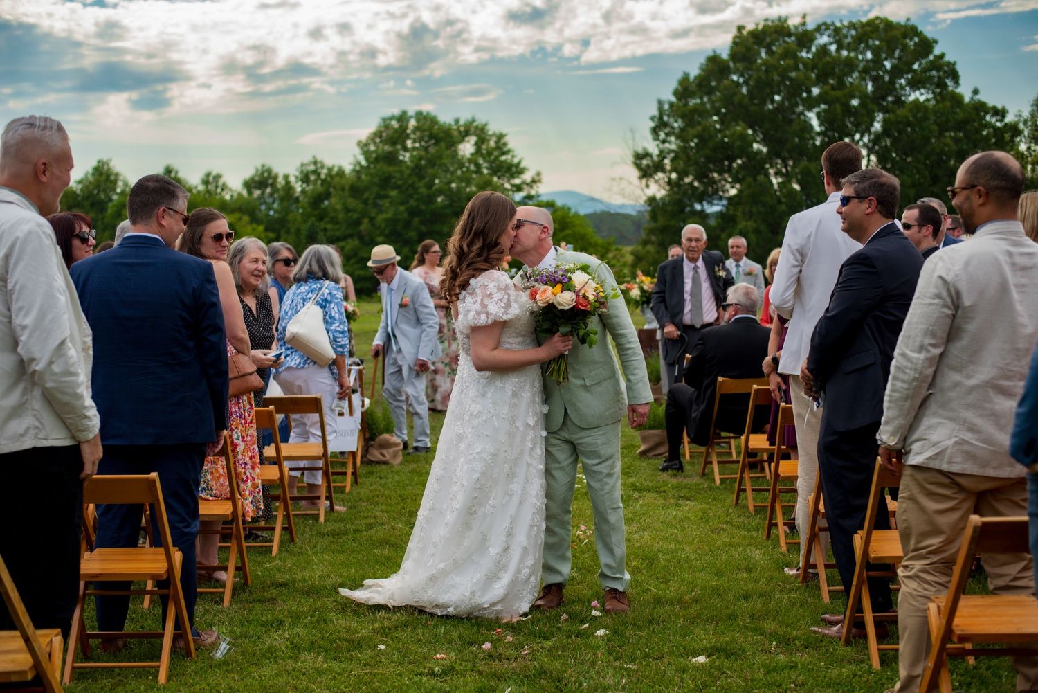 Bride and groom kissing in the aisle on wedding day surrounded by family. Sunny mountain background.