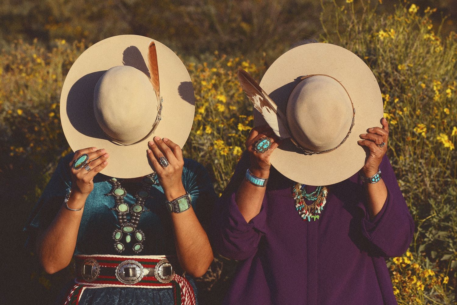 Two women in turquoise jewelry and traditional clothing hold wide-brimmed hats over their faces in a yellow wildflower field.