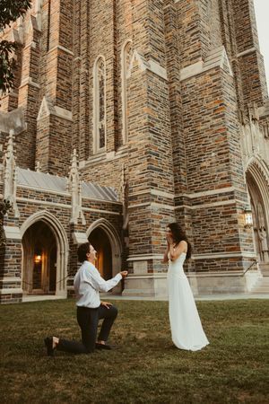 Groom proposing on one knee to bride in white gown outside Gothic stone church at dusk.