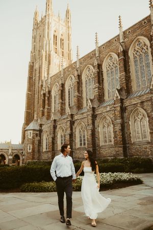 Couple holding hands walking in front of a Gothic cathedral in warm golden light, woman in white dress.