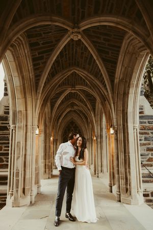 Couple embracing in gothic stone archway corridor, bride in white gown, groom in white shirt and dark pants.