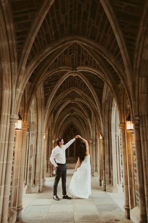 Couple dancing under Gothic stone arches in an elegant cathedral hallway, bride in flowing white gown.