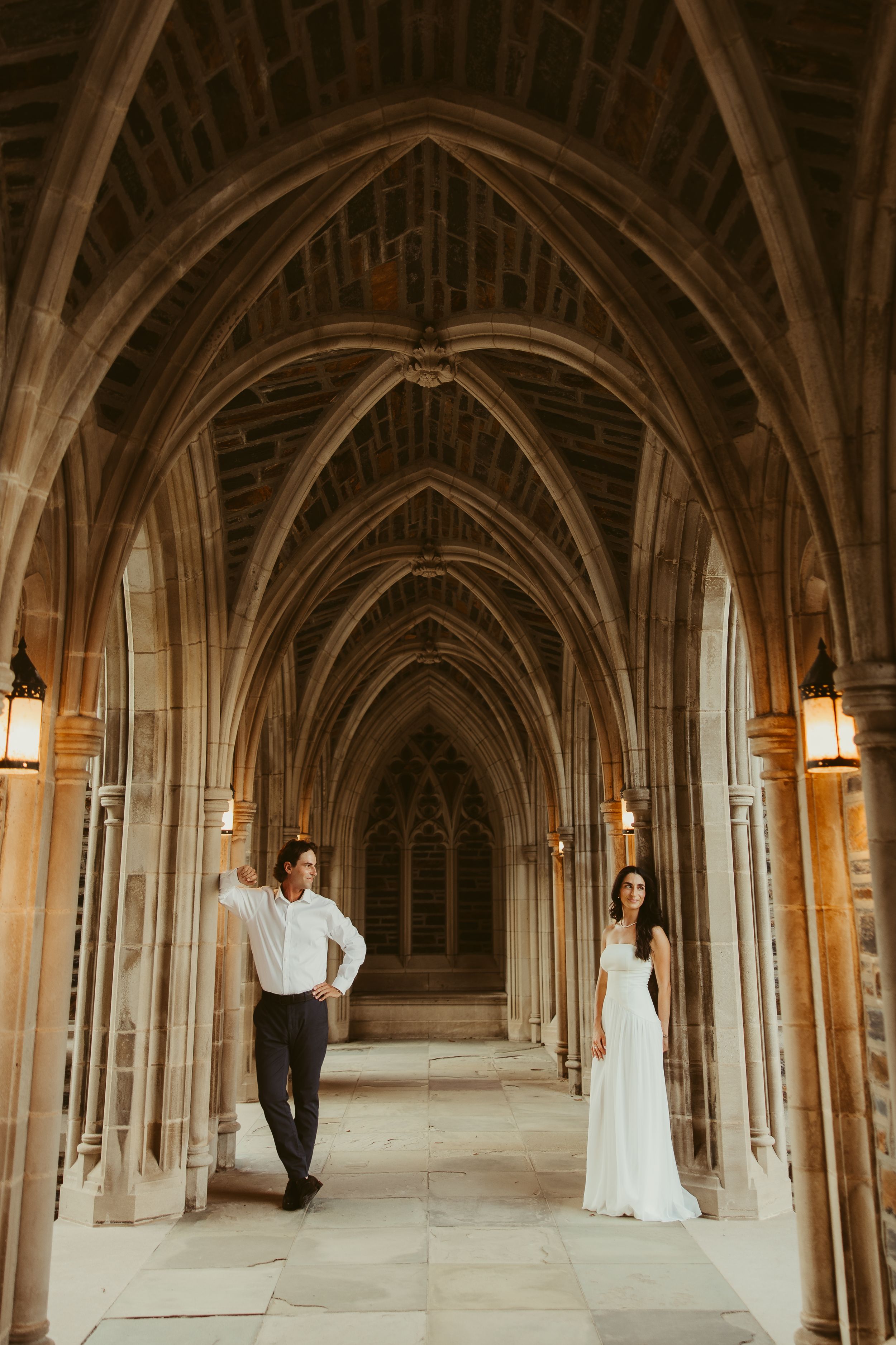 Couple poses in Gothic cathedral archway, bride in white gown, groom in white shirt and dark pants.
