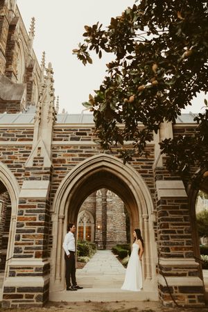Bride and groom stand beneath a Gothic stone archway at Duke Chapel, facing each other in wedding attire.