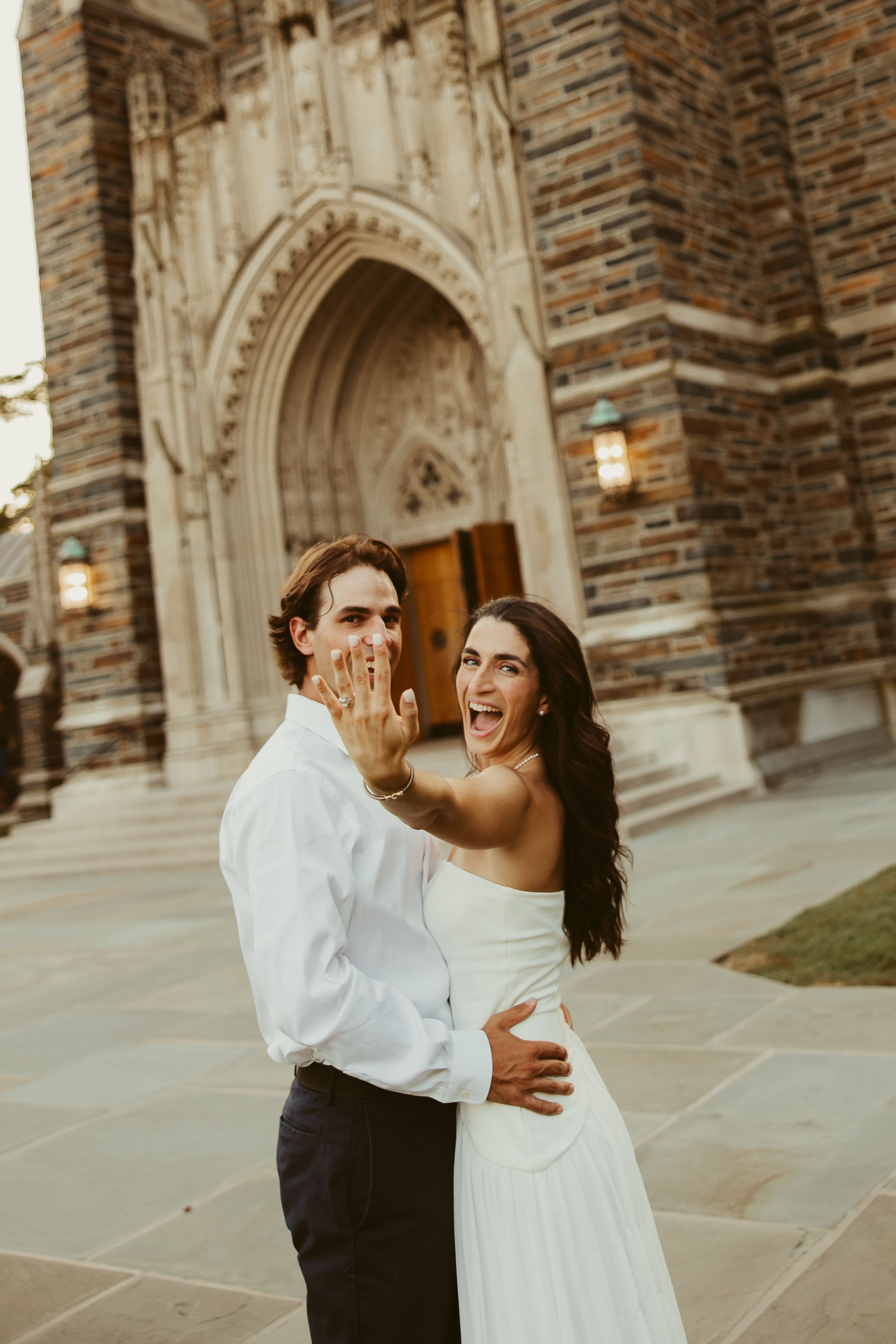 Excited bride shows engagement ring outside Gothic stone church, embraced by groom in white shirt.